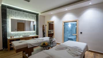 A modern hotel room with a marble-tiled vanity, two beds with towels, a small table with fruit and a glass, and a view of the city through a window.