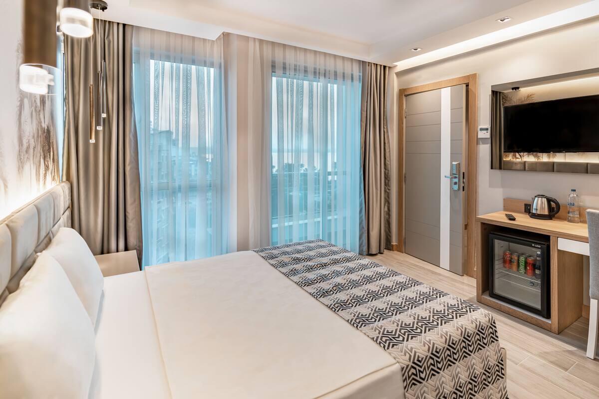 Une chambre d’hôtel moderne dotée d’un grand lit, d’un minibar, d’une télévision et offrant une vue sur la ville à travers de fines rideaux.