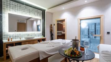 Een moderne hotelkamer met een grote spiegel, een houten bankje, een ronde tafel met een schaal fruit en wijn glazen, en uitzicht op de stad door een glazen deur.