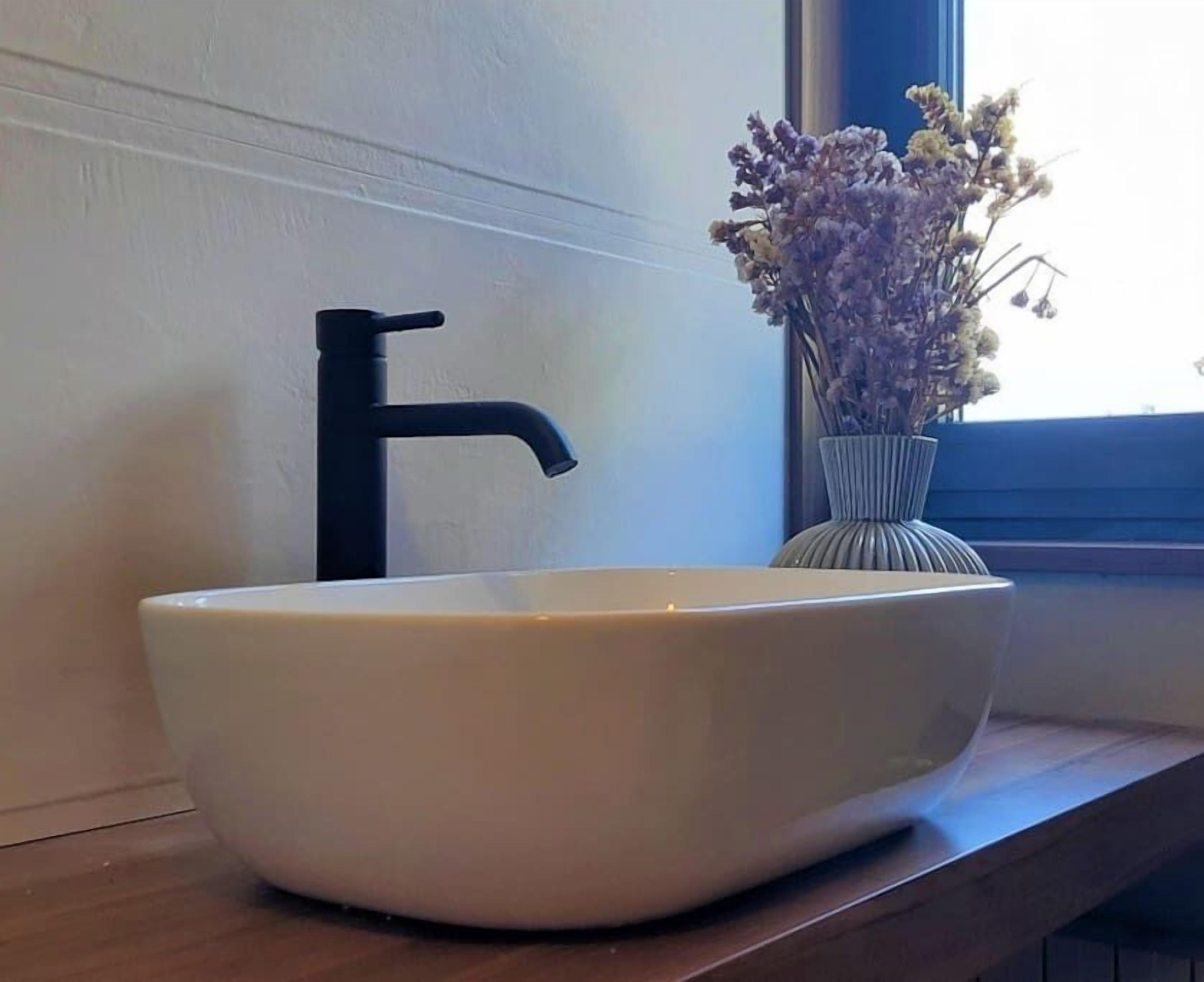 Lavabo bianco con rubinetto nero su un piano in legno, vicino a una finestra con un vaso di fiori secchi.