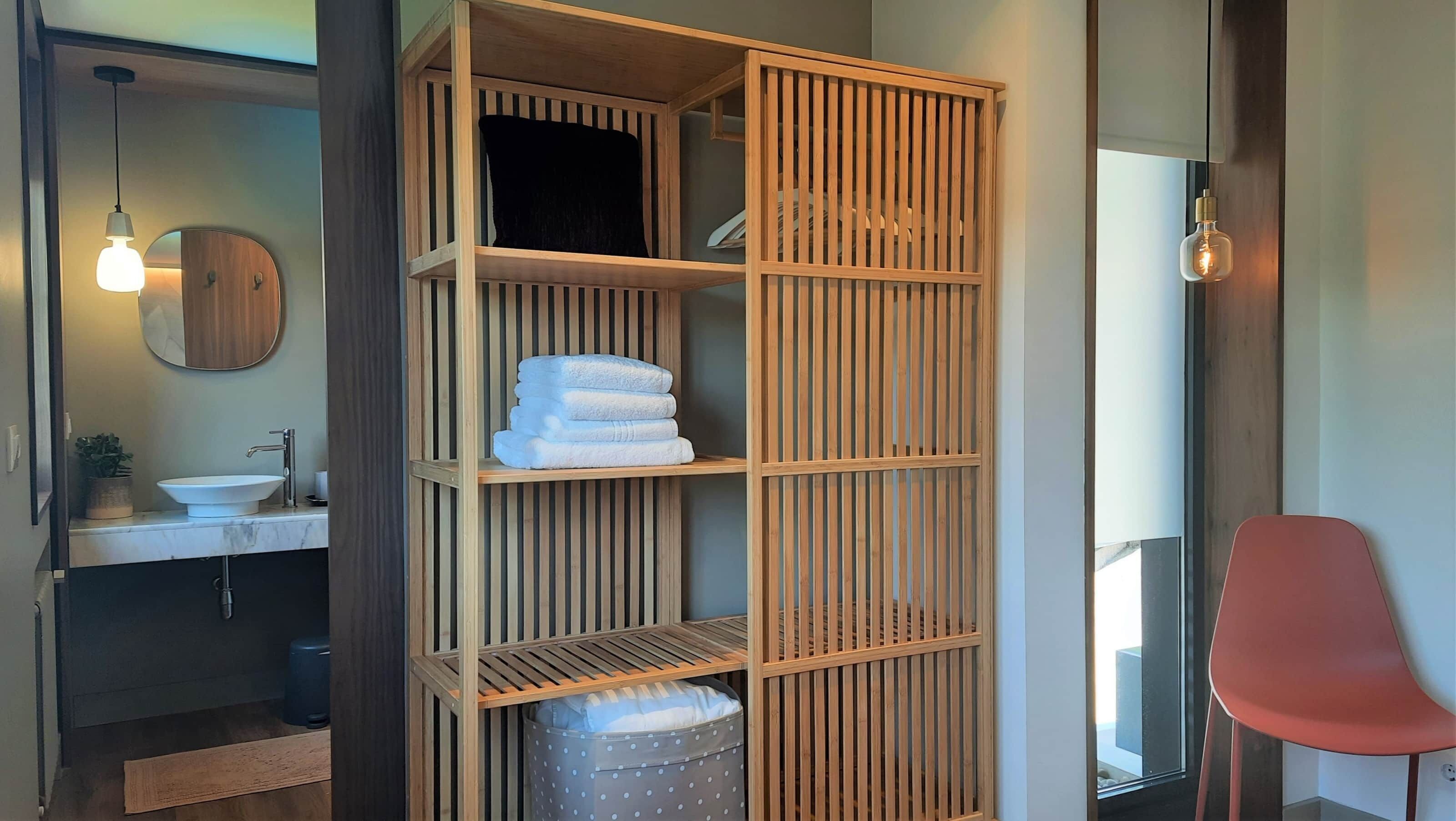 A modern bathroom with a wooden vanity, a sink, and a mirror. There is a red chair and a wooden shelf unit with towels.