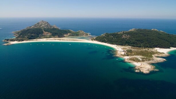 Vista aérea de una zona costera con una montaña destacada, una playa curvada y aguas azules cristalinas.