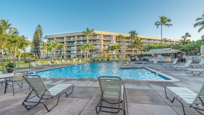 A hotel pool area with lounge chairs, palm trees, and a building with multiple balconies. - Oceanview suite nicely renovated B310 at Maui Sunset (Kihei)