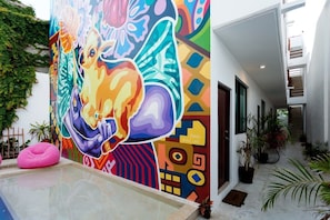 Um mural colorido numa parede de edifício, uma área de piscina e um corredor com plantas em vasos.
