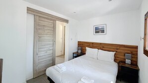Une chambre à coucher avec une tête de lit en bois, un lit recouvert de draps blancs, une table de chevet avec un haut-parleur, et une porte donnant sur une autre pièce.