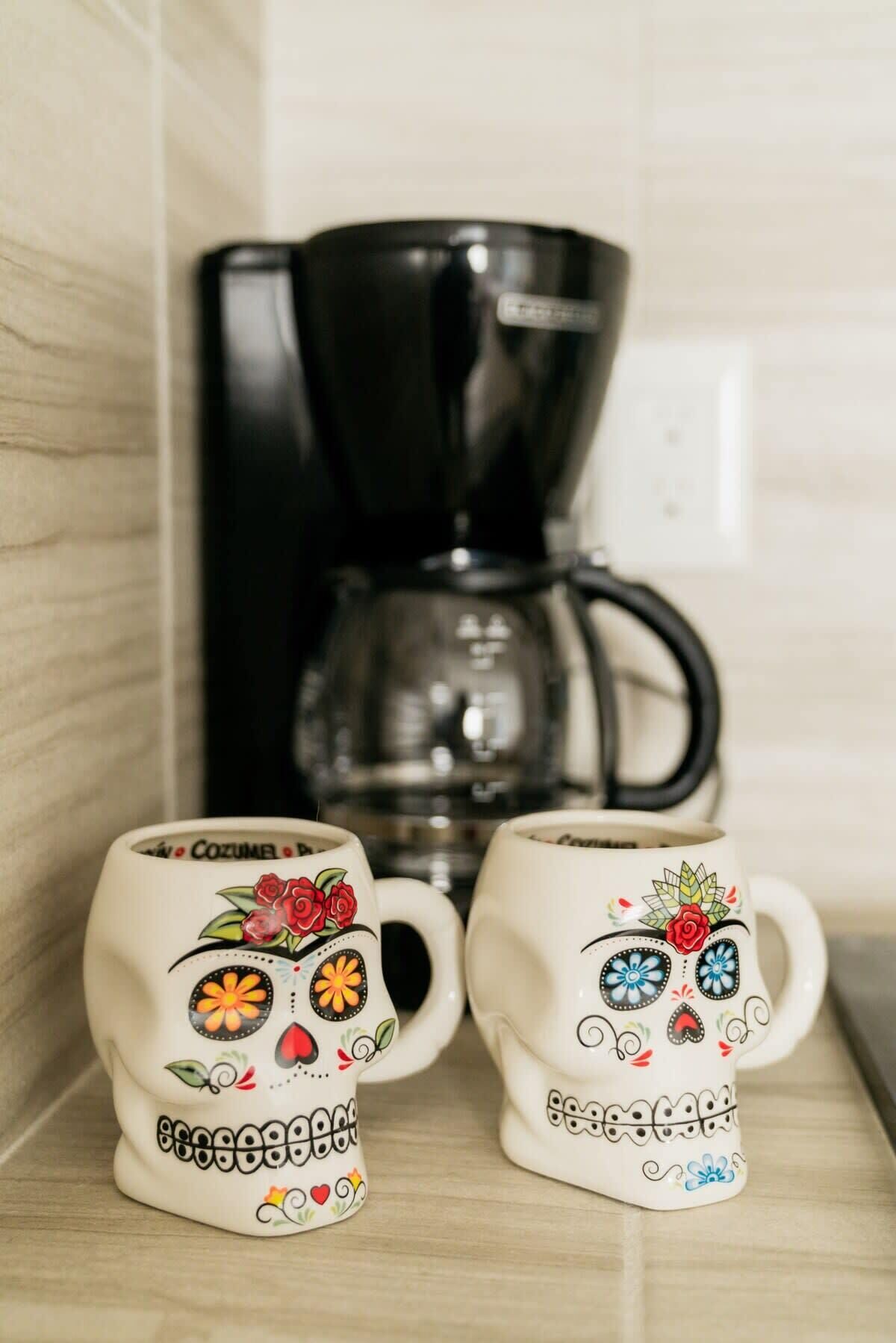 Two sugar skull mugs with floral designs on a shelf, a black coffee maker, and a tiled wall in the background.