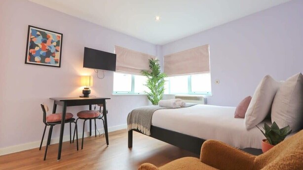 A bedroom with a bed, a desk with chairs, a TV, and a window with blinds. - Prince Street Studio 2 - Hopewell (Bristol)