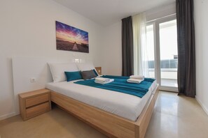 A modern hotel room with a large bed, a nightstand, a painting of a sunset, and a view of the outside through a window.