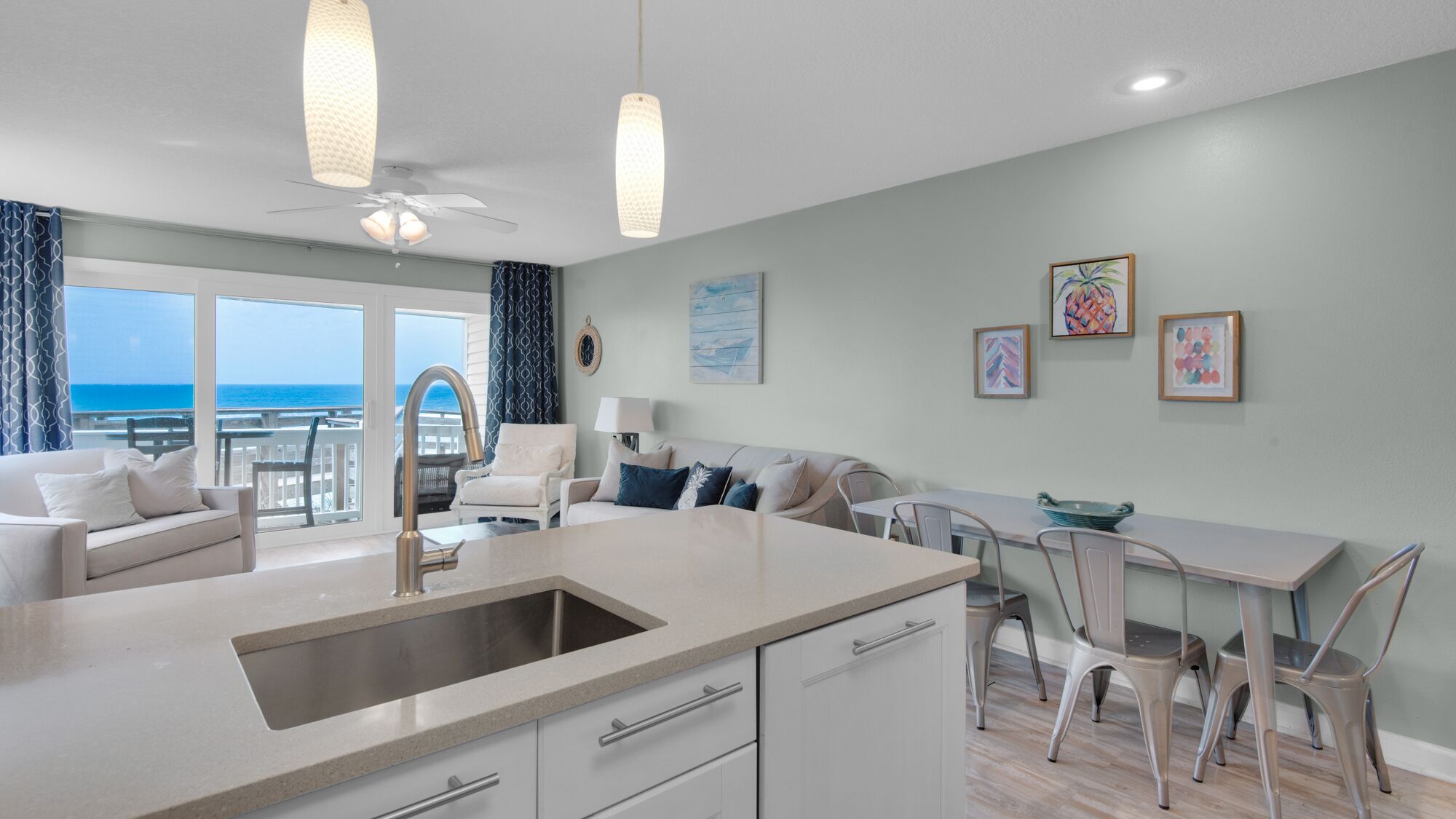 A modern kitchen with a large island, a dining area with a table and chairs, and a living area with a sofa and armchairs. There is a view of the ocean through large windows.