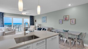 A modern kitchen with a large island, a dining area with a table and chairs, and a living area with a sofa and armchairs. There is a view of the ocean through large windows. - Gulf Front, Amazing Views, Beach Setup, Bare Feet Retreat + FreeTickets! (Seacrest)
