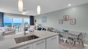 A modern kitchen with a large island, a dining area with a table and chairs, and a living area with a sofa and armchairs. There is a view of the ocean through large windows.
