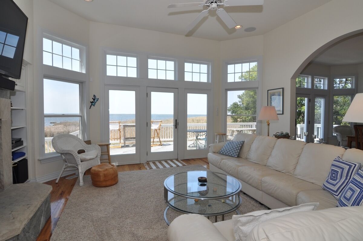 A spacious living room with a large glass coffee table, a beige sofa, and a white armchair. There are large windows offering a view of the ocean.