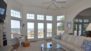 Ein geräumiges Wohnzimmer mit einem großen Glastisch, einem beigen Sofa und einem weißen Sessel. Große Fenster bieten Blick aufs Meer.