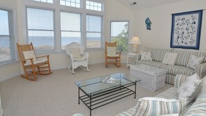 Ein geräumiges Wohnzimmer mit großen Fenstern, einem gestreiften Sofagarnitur, einem Glastisch und zwei Schaukelstühlen.