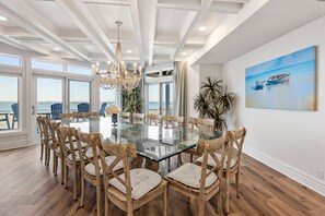 A dining area with a glass-top table surrounded by wooden chairs, large windows with a view of the beach, and a chandelier. - Bella Costa: Gourmet Kitchen, Pool, Spa, Elevator (Corolla)