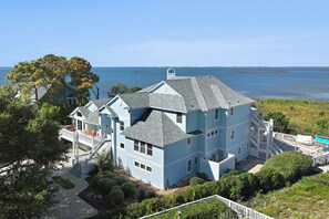 A large, light blue house with a covered porch and a pool, surrounded by greenery and overlooking a body of water. - Bella Costa: Gourmet Kitchen, Pool, Spa, Elevator (Corolla)