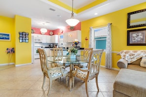 A dining area with a glass table surrounded by chairs, a kitchen in the background, and a living area with a sofa. - Mellow Yellow - Steps from the Beach - Fort Morgan - Signature Properties (Fort Morgan)