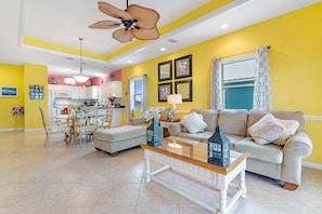 A bright living room with a yellow wall, a ceiling fan, a glass coffee table, and a sofa with cushions. - Mellow Yellow - Steps from the Beach - Fort Morgan - Signature Properties (Fort Morgan)