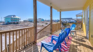 Una terraza de madera con sillas azules y una mesa, con vista a una playa y casas al fondo.
