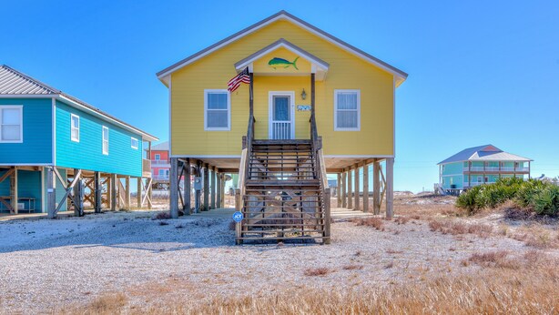 Una casa amarilla con una escalera de madera que sube a un porche, sostenida por pilotes sobre una playa arenosa.