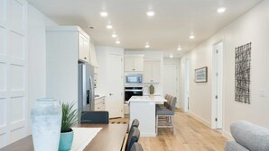 A modern kitchen with white cabinets, a built-in microwave, and a dining area with a wooden table and chairs. - Desert Color Lake House: Desert Color Lagoon + Beaches (St. George)