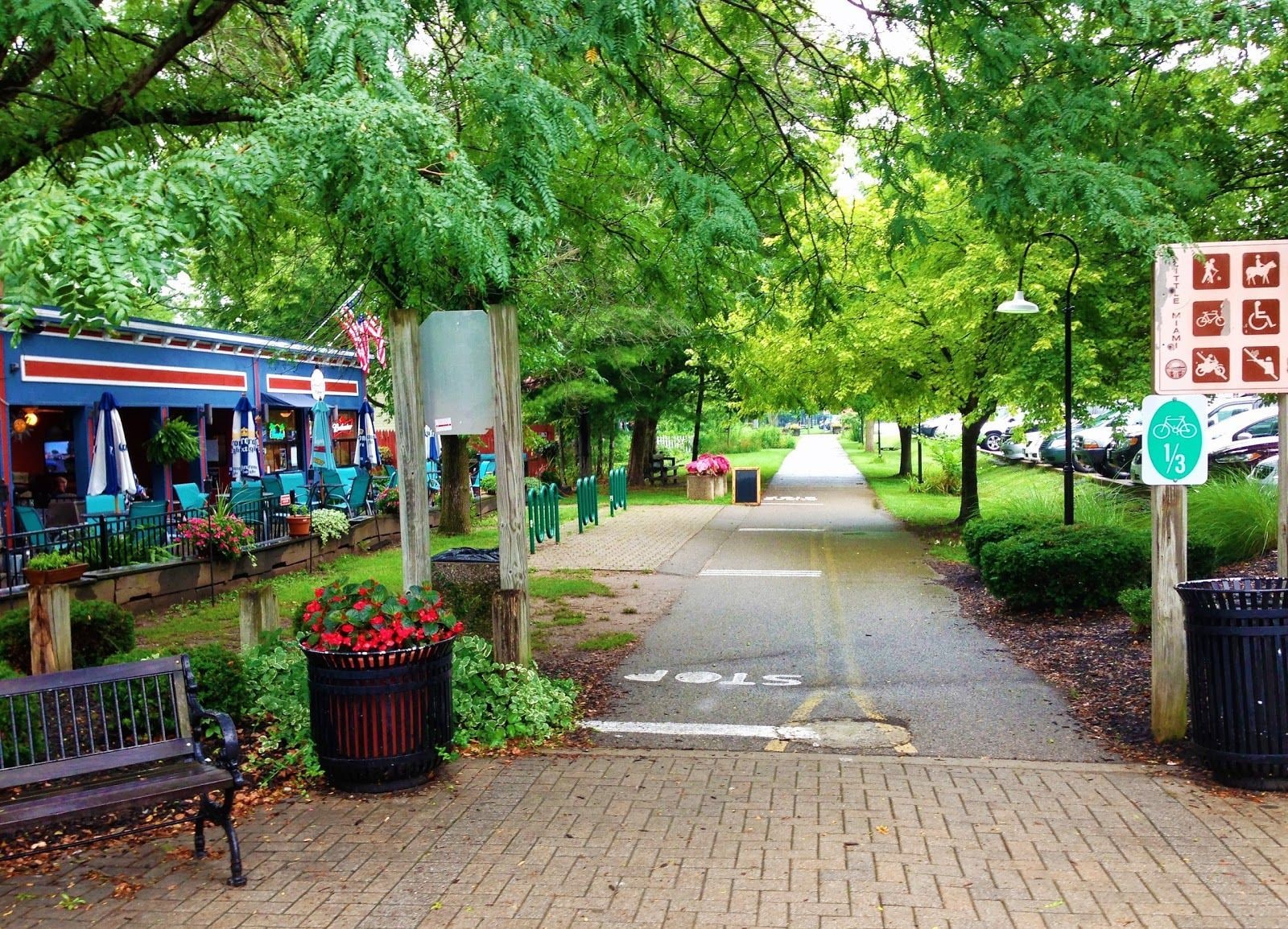 A pathway lined with benches and a trash bin, leading to a restaurant with outdoor seating.