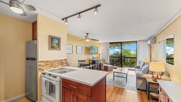 A modern kitchen with a central island, built-in oven, and refrigerator. There is a dining area with a table and chairs, and a balcony with a view of greenery. - Exceptional Maui Vista Condo With Ocean Views (Kihei)