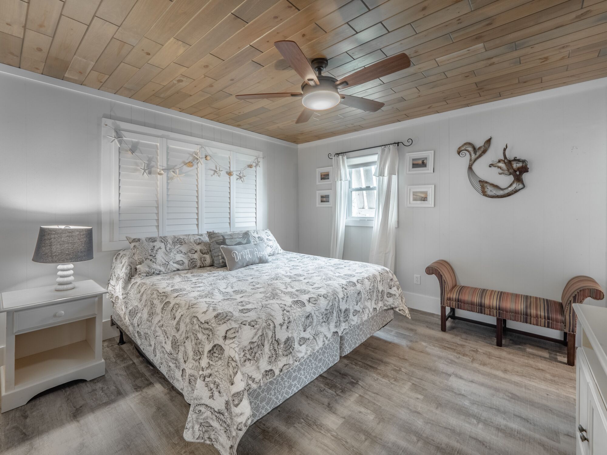 A bedroom with a bed, nightstand, bench, ceiling fan, and wall decor.