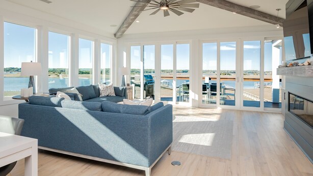 A spacious living room with a large blue sofa, a fireplace, and a view of the ocean. - Golden Hour: Luxurious Home with Pool Overlooking Lockwood Folly Inlet (Oak Island)