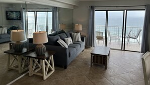 A modern living room with a sofa, two side tables with lamps, a small wooden table, and a balcony with chairs overlooking the ocean.