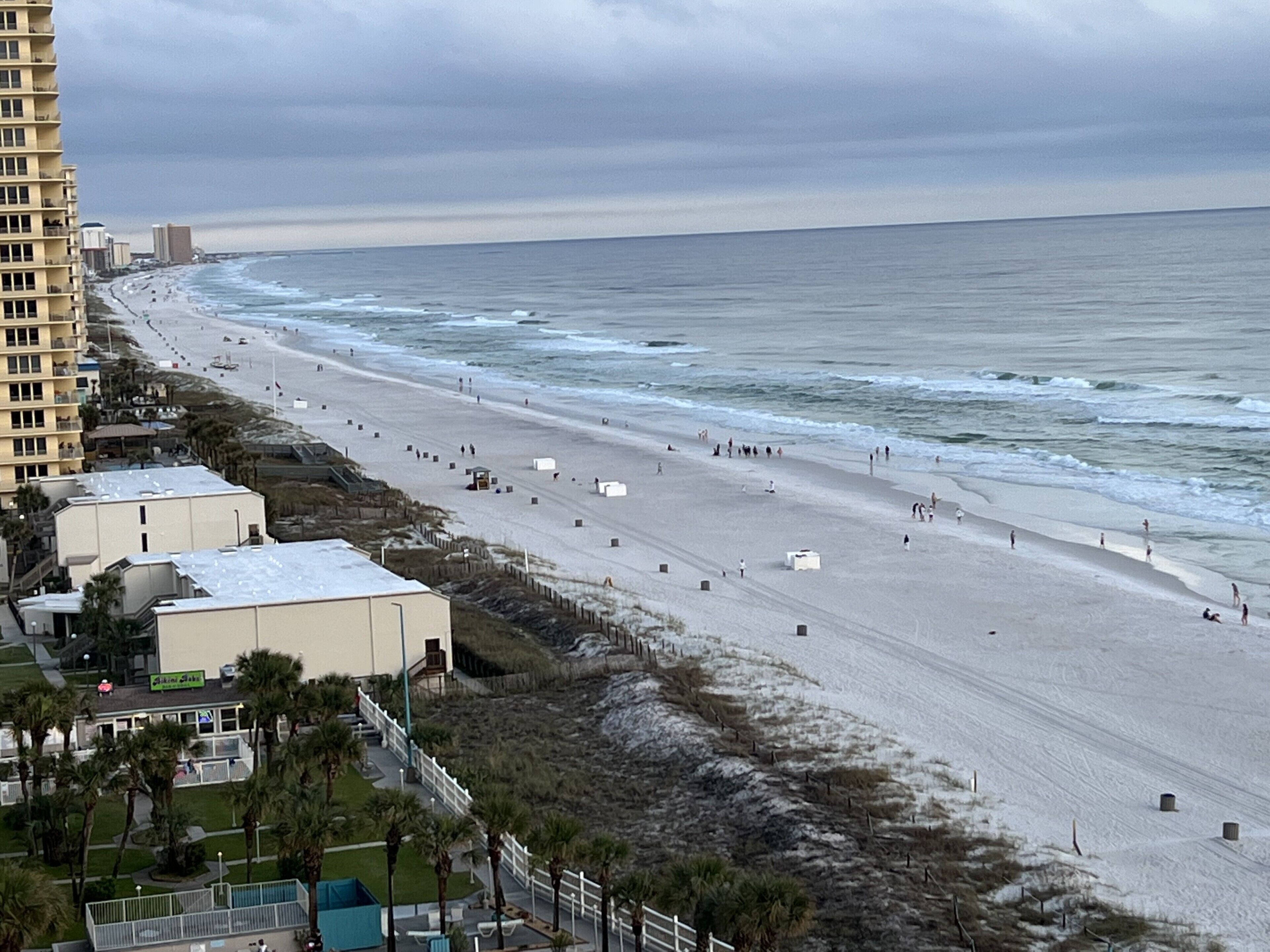 A beach with a long stretch of sand, a few buildings, and a tall hotel on the left.