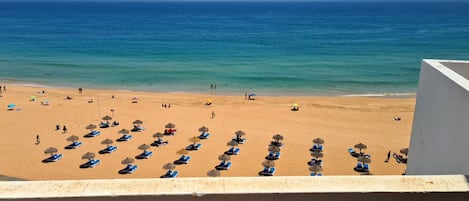 A beach with sun loungers and umbrellas, a clear view of the ocean, and a building in the foreground.