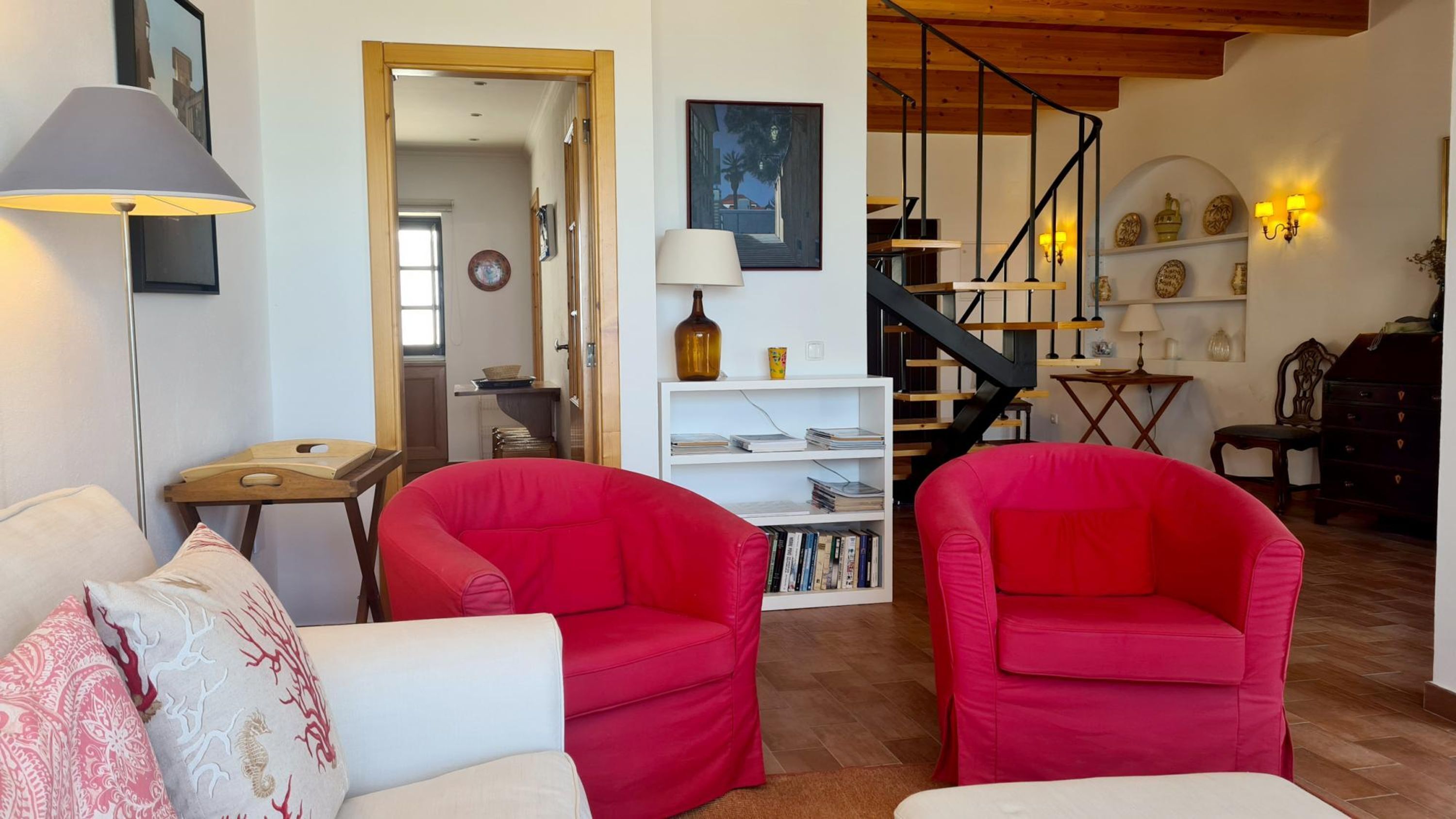 Una sala de estar con sillas rojas, un sofá blanco y una lámpara. Se observa una estantería y una escalera al fondo.