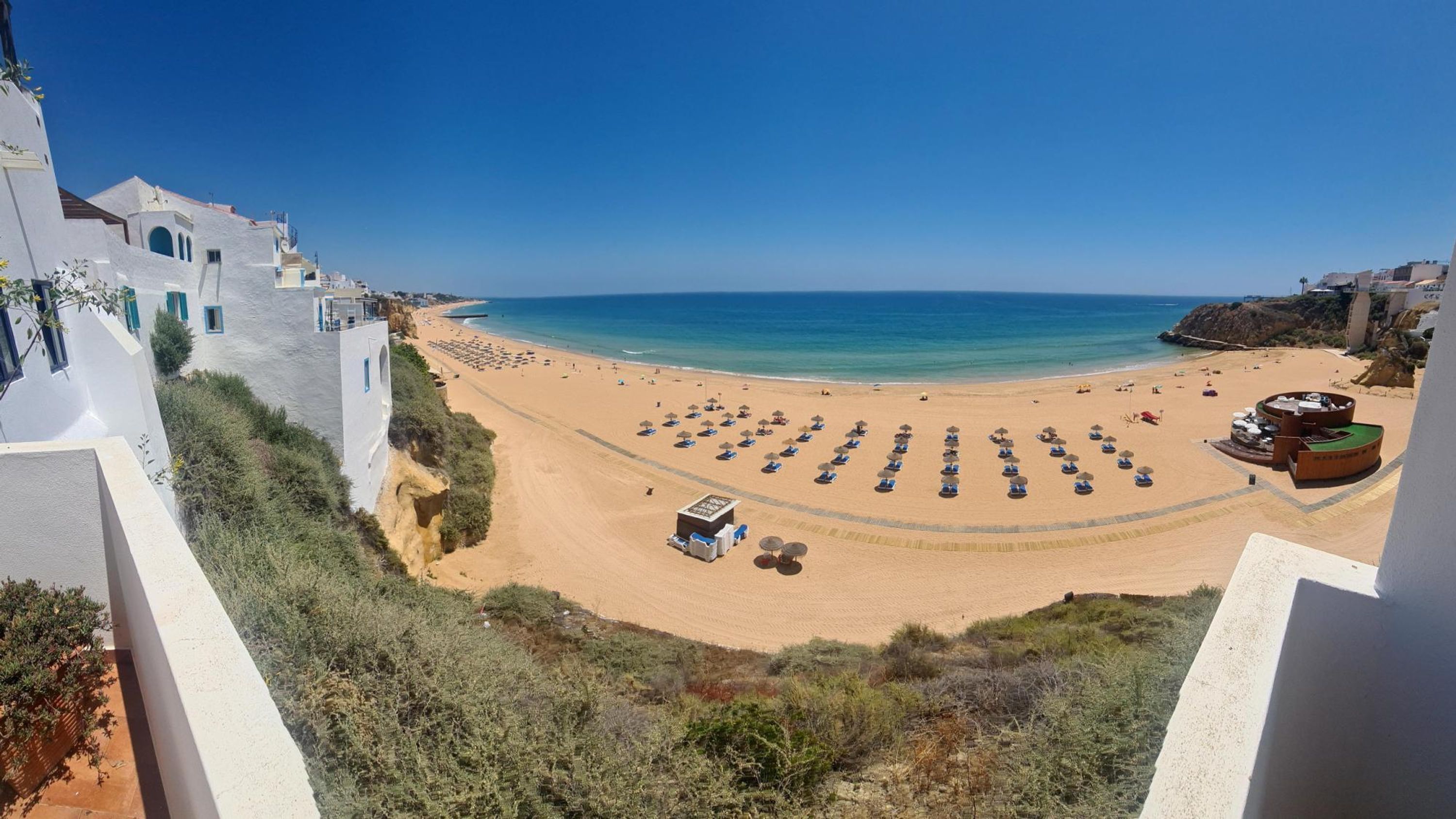 Une plage bordée de rangées de chaises longues et de parasols, un petit bâtiment doté d’un toit végétalisé et une vue dégagée sur la mer.