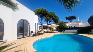 Uma área de piscina com piscina em curva, assentos externos e um edifício branco com janelas em arco.