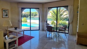 Ampla sala de estar com vista para a piscina, mesa de jantar com cadeiras e uma pequena mesa branca com luminária e revistas.