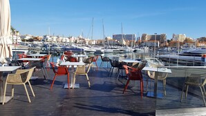 Uma marina com barcos atracados, área de estar ao ar livre com mesas e cadeiras, e edifícios ao fundo.