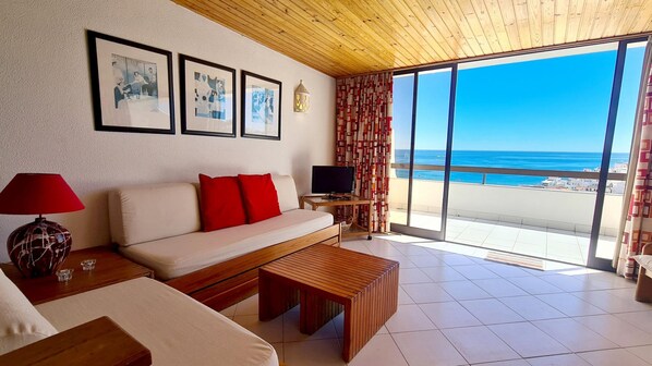 A modern living room with a sofa, red cushions, a TV, and a wooden coffee table. There are framed pictures on the wall, a lamp, and a view of the sea through large windows.