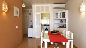 Een kleine keuken met een wasmachine, een eethoek met een rood tafelkleed en een wit kastje met glazen deuren.
