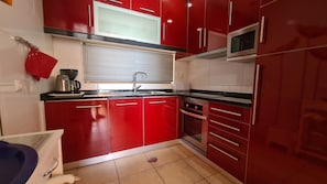 A kitchen with red cabinets, a microwave, a sink, and a window with blinds.