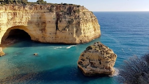A coastal cliff with a natural archway leading into the sea, surrounded by clear turquoise waters and rocky formations.