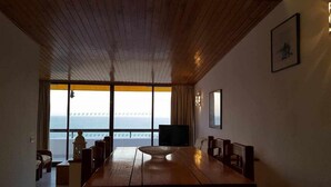 Un espace repas comprenant une grande table et des chaises en bois, un téléviseur et une vue sur l’océan grâce à de grandes fenêtres.