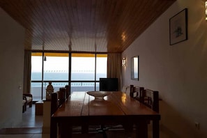A dining area with a large wooden table and chairs, a television, and a view of the ocean through large windows.