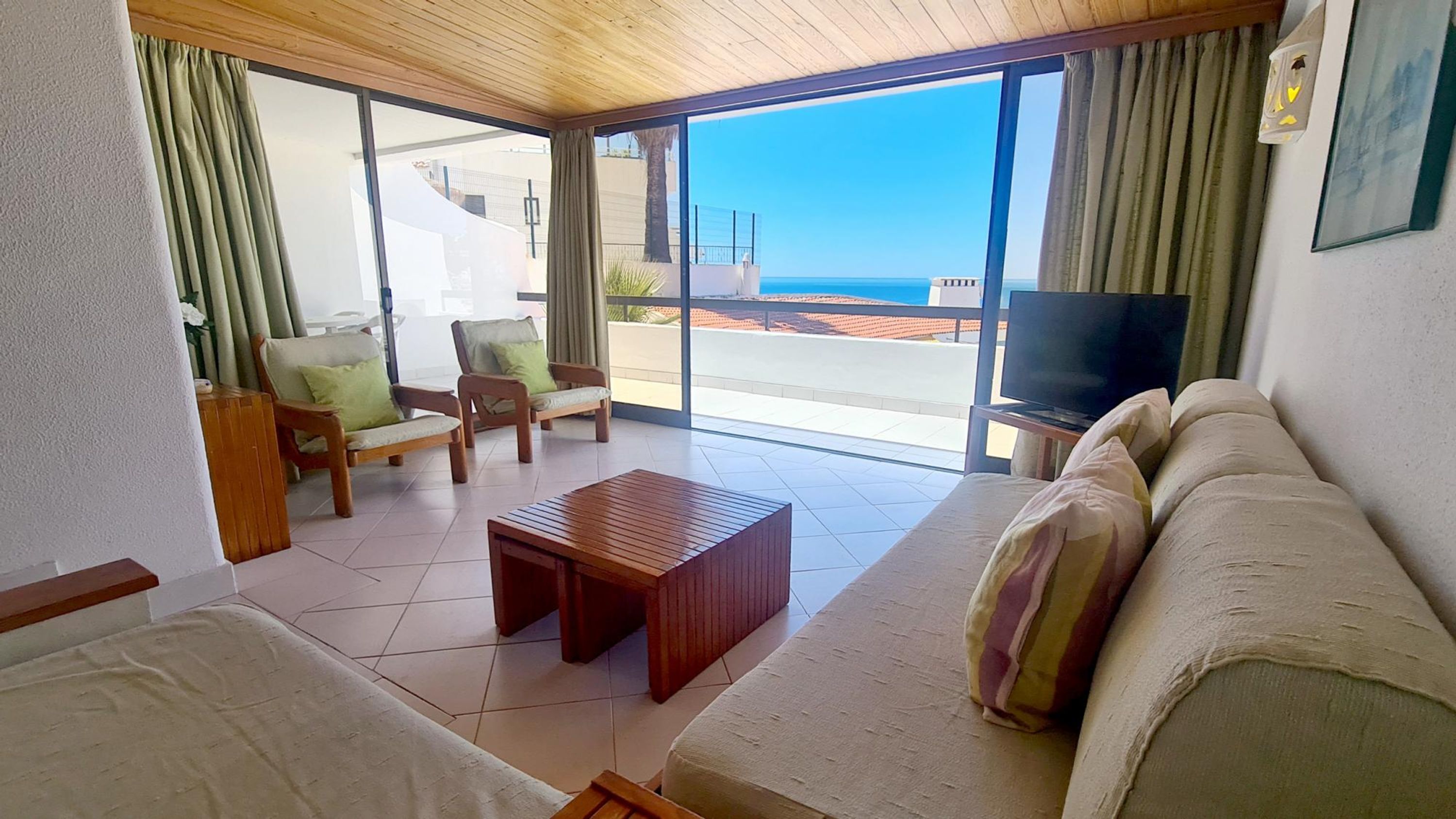 En stue med sofa, stole, et sofabord, et fjernsyn og en balkon med havudsigt.