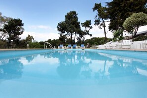Una piscina trasparente con una scaletta in metallo, circondata da poltroncine bianche e alberi.