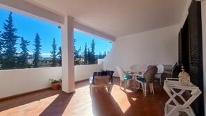A balcony with a wooden floor, white walls, and a view of trees and buildings. There is a table with chairs and a decorative lantern.