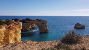 A coastal landscape with a prominent rock formation arching over the sea, a boat in the water, and a rocky shoreline. - Albufeira Balaia Mar e Sol by Rentals in Algarve (Albufeira)