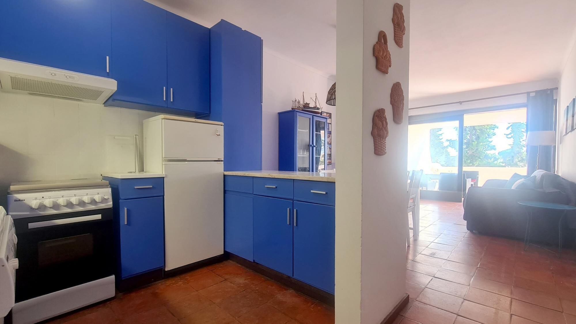 Cozinha com armários azuis, geladeira branca e fogão. Há objetos decorativos na parede e vista para o exterior por uma porta de vidro.