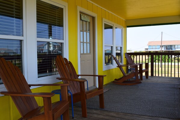 A porch with wooden chairs and a rocking chair, a yellow exterior wall, and a door with a sign.