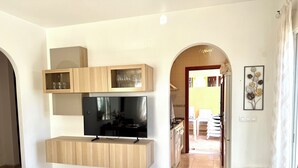 Uma sala de estar moderna com televisão de tela plana, armários de madeira, piso ladrilhado e vista para a área de jantar adjacente.